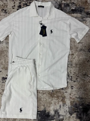Vit ribbad skjorta och shorts set - Snyggt vitt set med kortärmad skjorta och matchande shorts i ribbat tyg. Skjortan har knappar framtill och klassisk krage, båda plaggen har en broderad logga på bröstet och benet. Perfekt för en avslappnad och stilren look.