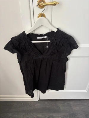 Lager 157 - Snygg svart blus från 157 i storlek XS. Blusen har korta ärmar med volangdetaljer och en djup v-ringning. Tillverkad i ett lätt tyg som ger en luftig känsla. Perfekt att matcha med jeans eller kjol för en trendig look.