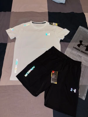Under Armour träningsset t-shirt & shorts - Snyggt träningsset från Under Armour med en vit t-shirt och svarta shorts. T-shirten har tryckta loggor och textdetaljer i regnbågsfärger, rund hals och kort ärm. Shortsen har elastisk midja och matchande logga. Perfekt för gym eller löpning.