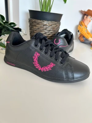 Svarta Fred Perry sneakers med rosa detaljer - Svarta sneakers från Fred Perry med rosa broderad logga på sidan och rosa detaljer på hälen. Skorna har lågt skaft, svarta skosnören och ovandel i läder med perforerade partier för extra stil. Perfekta för dig som vill ha en clean och sportig look.
