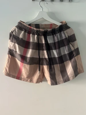 Burbbery shorts  - NFC SCAN fungerar.Snygga shorts med klassiskt rutmönster i beige, svart, vitt och rött. De har elastisk midja med svart dragsko och sidofickor. Materialet ser ut att vara lätt och bekvämt, perfekt för sommaren. Passar dig som gillar en trendig och avslappnad stil.
