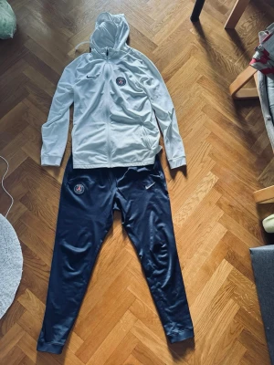 PSG Nike hoodie & byxor - Säljer ett PSG träningsset från Nike med vit hoodie och marinblå byxor. Hoodien har dragkedja, huva och PSG-logga på bröstet. Byxorna är smala, har snörning i midjan och PSG-märke på benet. 