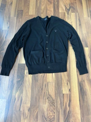 Svart cardigan från GANT - Klassisk svart cardigan från GANT med knäppning framtill och två fickor. Tillverkad i mjukt material och har ribbade muddar vid ärmslut och nederkant. Diskret GANT-logga broderad på bröstet. Perfekt för lager på lager och enkel att matcha.