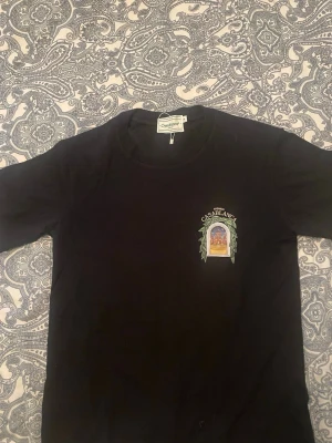Svart t-shirt från Casablanca - Snygg svart t-shirt från Casablanca med tryck på bröstet som föreställer ett fönster med gröna blad och texten 'Avenida Casablanca'. Klassisk passform och rund hals, perfekt till jeans eller shorts.