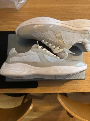 Gråa Prada cups - Snygga sneakers från Prada i grått och vitt med blank finish och meshdetaljer. Skorna har vita snören, tjock sula och röd logga på plösen. Perfekta för dig som gillar en clean och modern look.