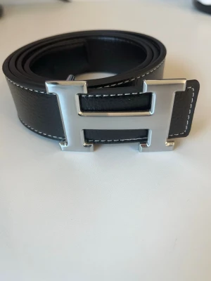Svart läderbälte med H-spänne - Snyggt svart bälte i läder med ett stort, silverfärgat H-format spänne. Bältet har vita kontrastsömmar längs kanterna som ger en clean och exklusiv look. Perfekt accessoar för att lyfta vilken outfit som helst.