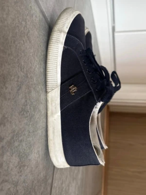 Mörkblå polo ralph lauren låga sneakers  - Snygga mörkblå sneakers  med vita gummisulor och matchande mörkblå skosnören. På sidan finns en liten guldfärgad logga som detalj.( polo ) Perfekta för en avslappnad och stilren look.