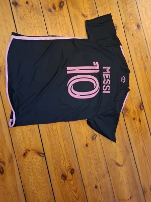 Svart Messi fotbollströja - Säljer en svart fotbollströja med rosa detaljer och trycket 'Messi 10' på ryggen. Tröjan har korta ärmar och är tillverkad i ett lätt och andningsbart material, perfekt för träning eller match.