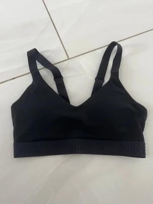 Svart sport-bh från Nike - Snygg svart sport-bh från Nike med justerbara axelband och bred resår med Nike-logga runt om. Tillverkad i ett mjukt och stretchigt material som ger bra stöd vid träning. Perfekt för gymmet eller andra sportaktiviteter.