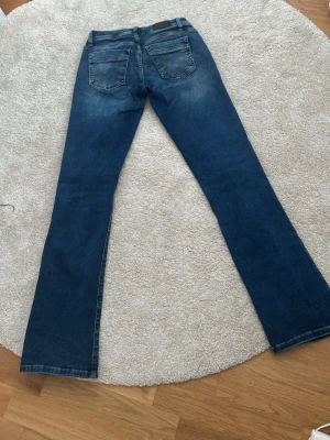 Blå bootcut jeans med slitningar - Snygga blå bootcut jeans med lätt slitna detaljer framtill och klassisk femficksdesign. Jeansen har en något utsvängd passform nertill och är gjorda i denim. Perfekta för en avslappnad och trendig look. Storlek 26/32