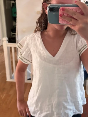 Vit blus med v-ringning och detaljer - Säljer en luftig vit blus med v-ringning och korta ärmar. Blusen har gröna broderade detaljer längs ärmsluten