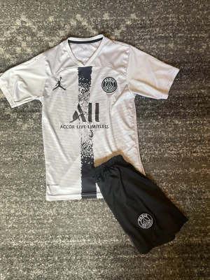 PSG matchställ i vitt och svart - Säljer ett snyggt PSG matchställ med grå tröja och svart lodrätt mönster samt svarta shorts. Tröjan har PSG-logga, Jordan-logga och sponsortryck framtill. Shortsen har elastisk midja och PSG-emblem. Perfekt för fotbollsträning eller match. Storlek 152, messi på ryggen med nummer 10