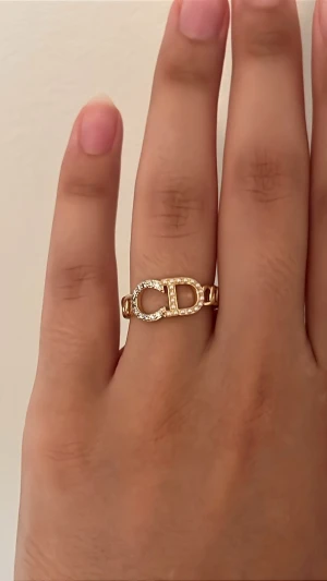 Guldfärgad ring med stenar och pärlor - Snygg guldfärgad ring med en kedjeformad design och en stor CD-detalj framtill, dekorerad med gnistrande små stenar. Ringen har en modern och trendig look som sticker ut och passar perfekt för dig som gillar statement-smycken.