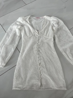 Vit kort klänning med broderi och knappar - Superfin vit kort klänning med broderade blommor, v-ringning och långa puffärmar. Klänningen har dekorativa knappar framtill och är tillverkad i ett lätt tyg som känns luftigt och fräscht. Perfekt för sommarens alla tillfällen! Endast använd 1 gång