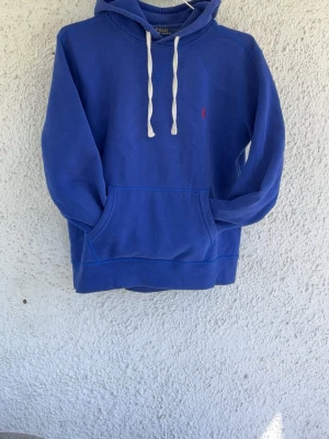Blå hoodie från Polo Ralph Lauren - Blå hoodie med vit dragsko och klassisk känguruficka framtill. Liten röd Polo-logga broderad på bröstet. Mjuk bomullskänsla och avslappnad passform, perfekt för en chill look.
