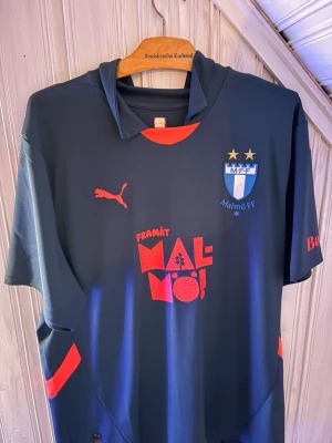 Malmö FF matchtröja Puma - Malmö FF matchtröja från Puma. Tröjan har klubbmärke, två stjärnor, och texten 'Framåt Malmö!' på bröstet. Boozt-logga på ärmen och klassisk sportig passform i lätt funktionsmaterial.