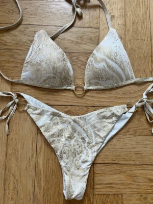 Vit och guld bikini med ringdetaljer - Säljer en snygg bikini i vitt och guld med marmorerat mönster. Överdelen har trekantskupor och knyt i nacken, medan underdelen har knyt i sidorna och guldfärgade ringdetaljer. Perfekt för sommarens strandhäng!