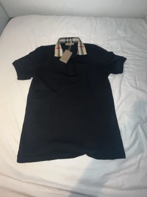 Svart Burberry pike - Snygg svart pikétröja från Burberry med klassisk rutig krage i beige, svart och rött. Helt ny. Endast testad. Strolek L. Passar även M. Pris kan diskuteras!