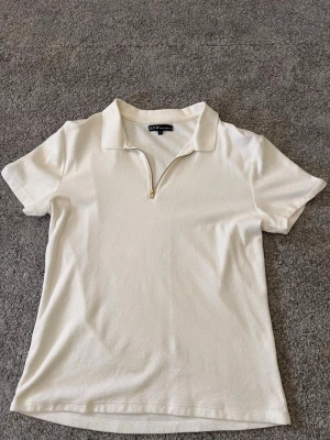 Vit kortärmad polotopp från SCW - Säljer en vit kortärmad polotopp från SCW med krage och guldfärgad dragkedja framtill. Toppen har en clean och enkel look som passar till det mesta. Materialet känns mjukt och bekvämt mot huden.