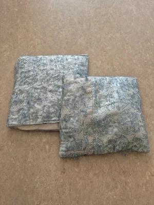 Kudde och kuddfodral - Säljer två kuddfodral med kudde silver tyg med ett broderat mönster. Fodralen har en fyrkantig form och tyget har en glansig yta med struktur. Perfekt för att ge soffan eller sängen en lyxig touch. 10kr för allt.