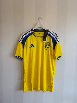 Svensk landslagströja Adidas gul - Gul svensk landslagströja från Adidas med blå detaljer och vita ränder på axlarna. Tröjan har rund halsringning, korta ärmar och Sveriges landslagsemblem på bröstet. Perfekt för fotbollsfans som vill stötta Sverige.