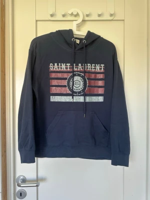 Mörkblå hoodie med tryck - De är stl M men sitter bra om man har stl S. Snygg mörkblå hoodie med huva och dragsnören. Framtill finns ett stort tryck med texten 'Saint Laurent' och röda samt vita ränder. Klassisk känguruficka och ribbade muddar vid ärmar och nederkant. Perfekt för en avslappnad streetwear-look.