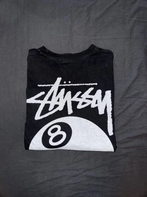 Svart Stüssy t-shirt med 8-ball print - Svart t-shirt från Stüssy med stort vitt 8-ball tryck och logga på ryggen. Klassisk streetwear-look med korta ärmar och rund hals. Perfekt för dig som gillar grafiska prints och vill sticka ut. Tillverkad i mjuk bomull för skön känsla.