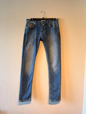 Blå raka Morris jeans Storlek 29/32 - Snygga blå Morris jeans med rak passform och guldfärgad broderad lilja på myntfickan. Jeansen har klassisk femficksdesign, guldfärgade nitar och kontrastsömmar. Perfekta för dig som gillar en enkel men ändå unik look. Storlek 29/32