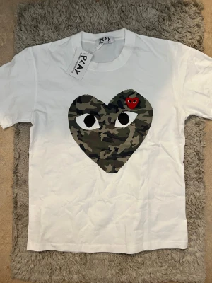 Vit t-shirt med camo hjärta från Play - Vit t-shirt med stort hjärta i camouflage-mönster och ögon på bröstet, samt ett litet rött hjärta med ögon upptill. Klassisk rund hals och korta ärmar. Perfekt för dig som gillar streetstyle och unika prints.