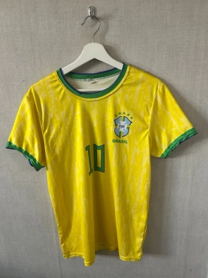 Brasilien tröja Pelé nummer 10 - Gul Brasilien fotbollströja med gröna detaljer, nummer 10 och Pelé på ryggen. CBF-emblem på bröstet och klassisk rund hals med grönt och blått. Perfekt för dig som älskar fotboll och vill hylla en legend.
