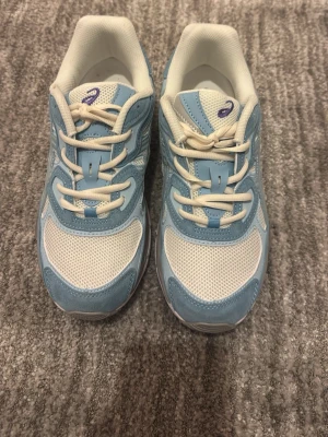 Blåvita sneakers med mesh och mocka - Snygga sneakers från Asics i ljusblått och vitt. Ovandelen är i mesh och mockaliknande material med vita snören och detaljer. Skorna har en sportig vibe och är perfekta för dig som gillar stilrena och bekväma sneakers.