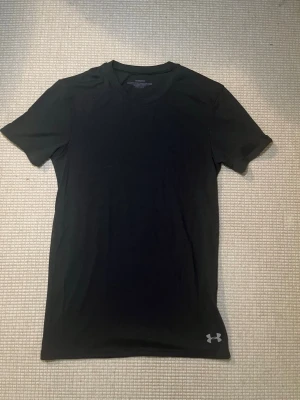 Svart träningströja från Under Armour - Säljer en svart, kortärmad träningströja från Under Armour. Tröjan har en tight passform och är tillverkad i ett lätt och snabbtorkande material, perfekt för intensiva träningspass. Under Armour-logga finns både fram och bak.