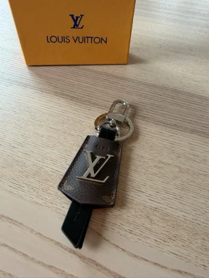  Vuitton nyckelring brun monogram - Snygg nyckelring från  Vuitton i brunt monogramläder med silverfärgad LV-logga framtill. Har metallring och karbinhake i silver för enkel fastsättning. Perfekt accessoar för att ge väskan eller nycklarna en lyxig touch.