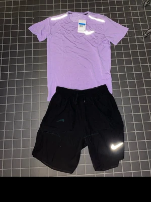 Lila tränings-tshirt & svarta Nike shorts - Säljer ett set med en lila tränings-tshirt och ett par svarta Nike shorts. Shortsen har resårmidja med dragsko och en vit Nike-logga på benet. Perfekt för gymmet eller löprundan. Materialet är lätt och andas, vilket gör dem sköna att träna i.
