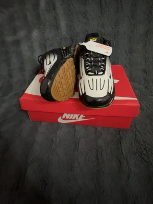 Nike svartvita sneakers med luftdämpning - Nike sneakers i svart och vitt med futuristisk design och synlig luftdämpning i sulan. Ovandelen har svarta och vita paneler med svarta detaljer och snörning. Yttersulan är brun och ger bra grepp. Perfekta för dig som gillar streetwear och sportig stil.