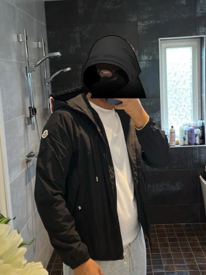 Svart Moncler vindjacka med huva helt ny  - Snygg svart vindjacka från Moncler med huva och dragkedja. Jackan har Moncler-logga på ärmen och detaljer i rött, vitt och blått på huvan. Perfekt för dig som vill ha en stilren och sportig look. Materialet är tunt och vindtätt.billigast på plick 
