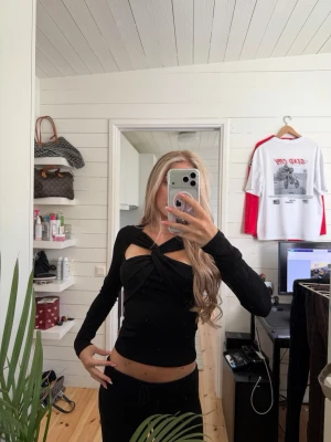 Svart långärmad topp med twist - Trendig svart långärmad topp med twistad detalj framtill och cut-out vid bröstet. Toppen är tight och croppad, vilket ger en snygg siluett. Perfekt för dig som vill ha en unik och stilren look.