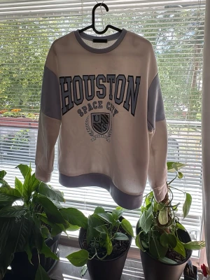 Vit och grå sweatshirt med Houston-tryck - Cool sweatshirt i vitt och grått med stort 'HOUSTON SPACE CITY' tryck på bröstet och US-emblem. Tröjan har kontrastfärgade muddar och partier på ärmarna samt rund hals. Perfekt för en avslappnad streetwear-look.