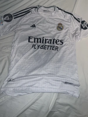Real Madrid matchtröja Mbappé #9 - Vit Real Madrid matchtröja med Mbappé och nummer 9 på ryggen. Tröjan har svarta detaljer, klubbmärke och Adidas-logga på bröstet samt sponsortryck. Tillverkad i lätt och ventilerande material med HEAT.RDY-teknologi. Perfekt för fotbollsträning eller match.