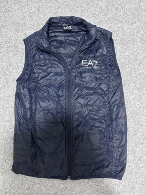Mörkblå dunväst från EA7 Armani - Snygg mörkblå dunväst från EA7 Emporio Armani med diskret glansig finish och broderad logga på bröstet. Västen har hög krage, dragkedja framtill och två sidofickor. Perfekt för lager-på-lager och streetwear-stil.