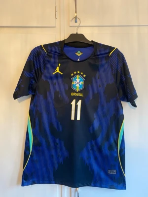 Blå Brasilien matchtröja Raphinha 11 - Snygg blå matchtröja för Brasilien med gula detaljer och nummer 11, Raphinha på ryggen. Tröjan har korta ärmar, rund hals och officiellt CBF-märke på bröstet. Tillverkad i lätt och ventilerande material, perfekt för fotboll eller träning.