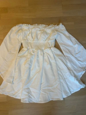 Vit offshoulder kortklänning - Söt vit kortklänning med offshoulder-design och långa, vida ärmar. Klänningen har smock i midjan för en markerad siluett och volangdetaljer vid halsringningen. Tillverkad i ett lätt tyg som ger ett luftigt intryck. Miniklänning med diagonala vida ärmar 