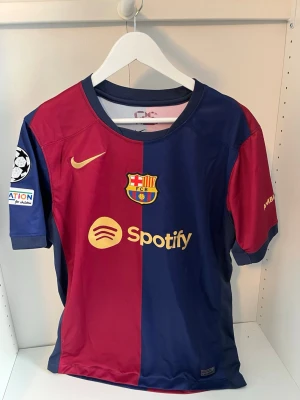FC Barcelona matchtröja Nike - Snygg FC Barcelona matchtröja från Nike i rött och marinblått med klubbmärke, Spotify-logga och Champions League-märke på ärmen. Tillverkad i lätt och ventilerande material, perfekt för fotboll eller träning. Ambilight tv-tryck på ena ärmen.