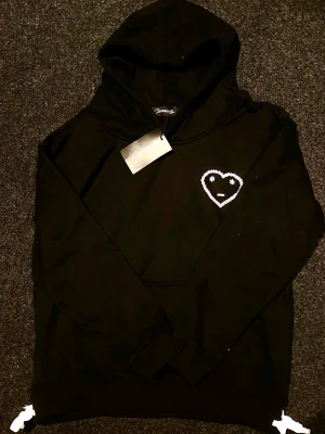 Svart Carsicko Hoodie - Cool svart hoodie från Carsicko med ett vitt hjärtmotiv och ansikte på bröstet. Den är helt ny, aldrig använd och kommer med tags. Perfekt för dig som gillar streetwear!                                                       🤝📦 Fraktar inom 24 timmar