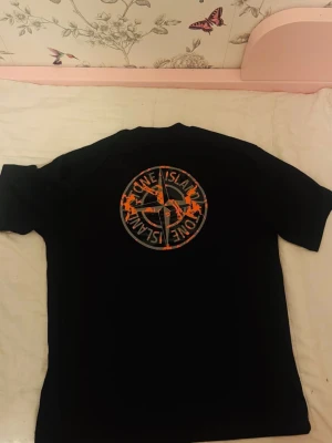 Svart Stone Island t-shirt med tryck - Svart t-shirt från Stone Island med orange logga på bröstet och stort tryck på ryggen. Klassisk passform och rund halsringning. Perfekt för dig som gillar streetwear och vill sticka ut med en snygg detalj på ryggen.