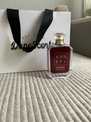 Kyneti Vaniljparfym med lyxig flaska - Kyneti parfym med doft av vanilj i en elegant, mörkröd glasflaska med guldiga detaljer och en kristalliknande kork. Flaskan har en modern, kantig form och ger ett exklusivt intryck. Perfekt för dig som gillar unika och stilrena parfymer.