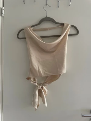 Champagnefärgad omlottopp med knyt - Säljer en champagnefärgad omlottopp i satin med snygg knytning i midjan. Toppen är ärmlös och har en draperad urringning som ger en elegant look. Perfekt för dig som vill ha något trendigt och stilrent i garderoben.