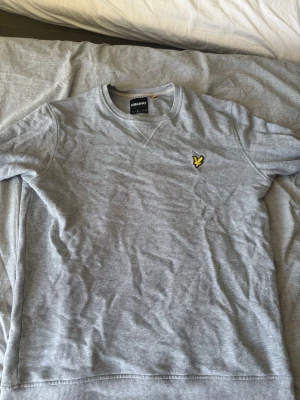 Grå sweatshirt från Lyle & Scott - Säljer en stilren grå sweatshirt från Lyle & Scott med klassisk rund halsringning och den ikoniska gula örnloggan broderad på bröstet. Tröjan har ribbade muddar vid ärmslut och nederkant. Perfekt för en avslappnad look.