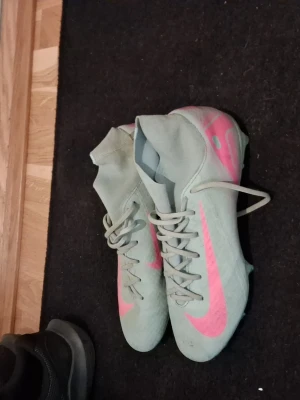 Turkosa Nike fotbollsskor med rosa detaljer - Säljer ett par turkosa Nike fotbollsskor med högt skaft och rosa swoosh på sidan. Skorna har snörning och dobbar för bra grepp på planen. Perfekta för dig som vill sticka ut på fotbollsplanen.