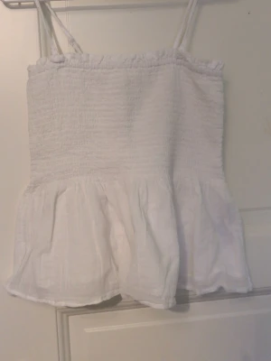 Vit smockad linnetopp Gina Tricot - Söt vit linnetopp från Gina Tricot med smockad överdel och tunna axelband. Toppen har volang nedtill och är perfekt för varma dagar. Enkel och stilren design som passar till det mesta.